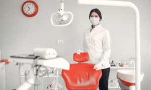Female dentist in white coat and mask holding red dental chair cushion in modern treatment room.
