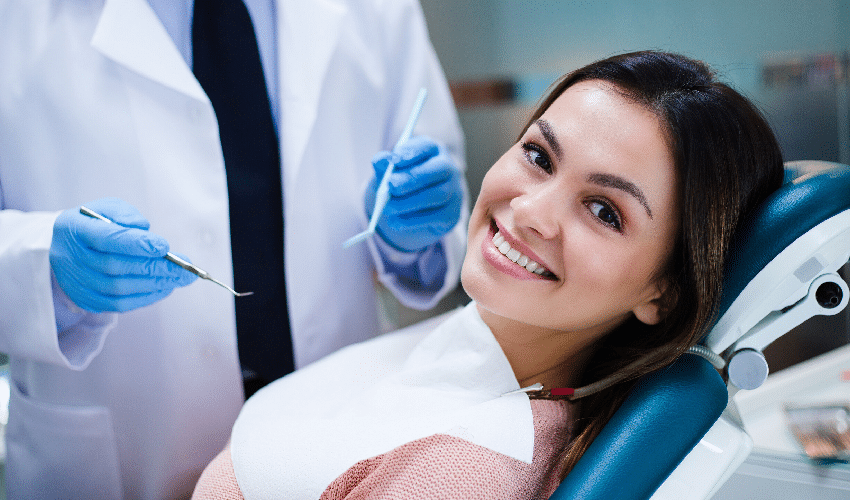 Smiling female patient in dental chair with dentist holding tools during examination.